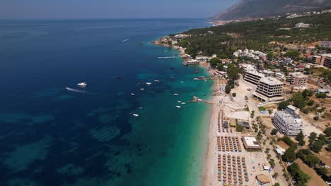 Tour-boats-floating-near-pier-at-beautiful-beach-of-Dhermi-in-Albania,-row-of-umbrellas-near-blue-turquoise-sea