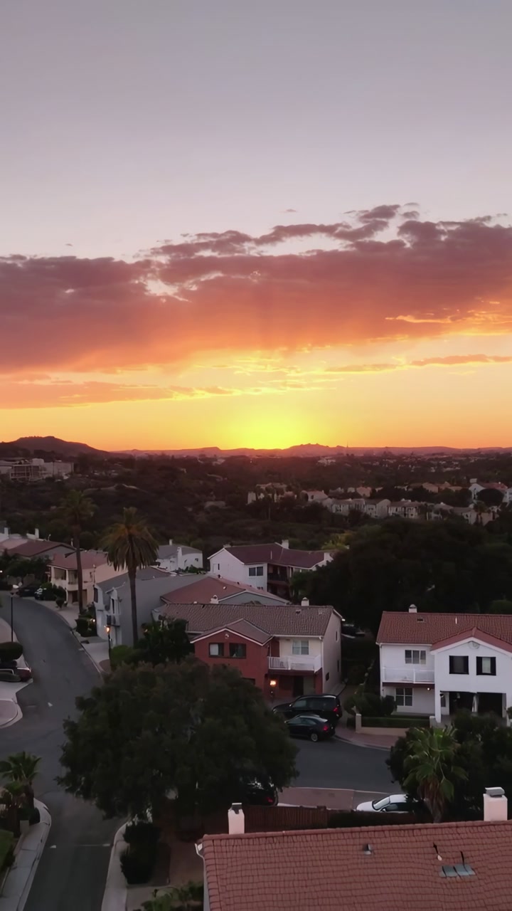 Vertical video: Sinking sun disk casting warm glow while camera tilting, revealing suburban roofs