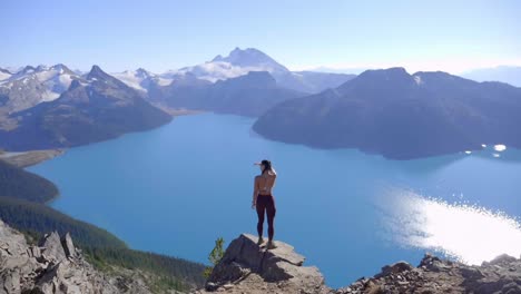 Mujer-Excursionista-De-Pie-Sobre-Una-Roca-Mirando-Por-Encima-De-Un-Lago-De-Montaña