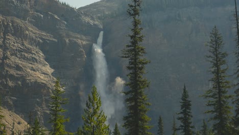 a cinematic and beautiful slow motion shot of huge pine trees and a the very impressive takakkaw waterfall in the background