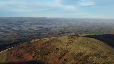 Early-sunlight-on-highland-mountain-peak-aerial-view-across-vast-frosty-idyllic-farmland-countryside-wilderness-pull-back-shot