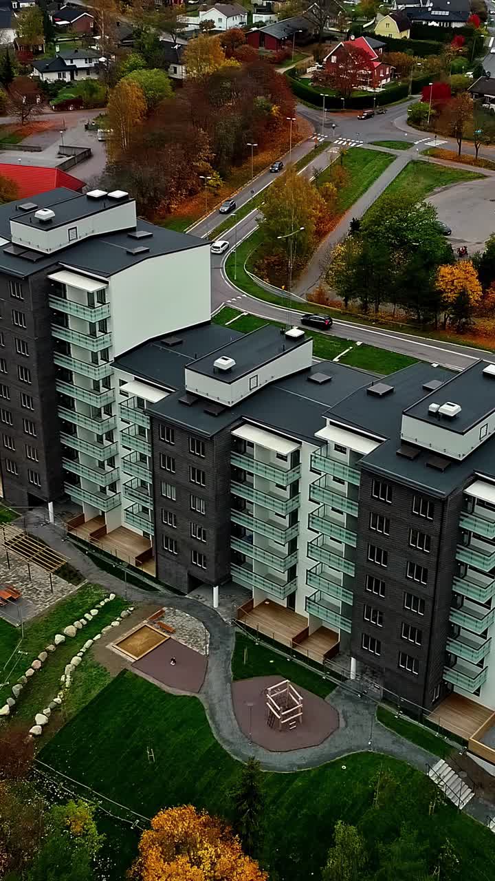 Una ripresa aerea in discesa verticale mostra un condominio moderno di nuova costruzione con un parco giochi, circondato da alberi autunnali colorati in un tranquillo sobborgo residenziale in Lettonia, immobiliare