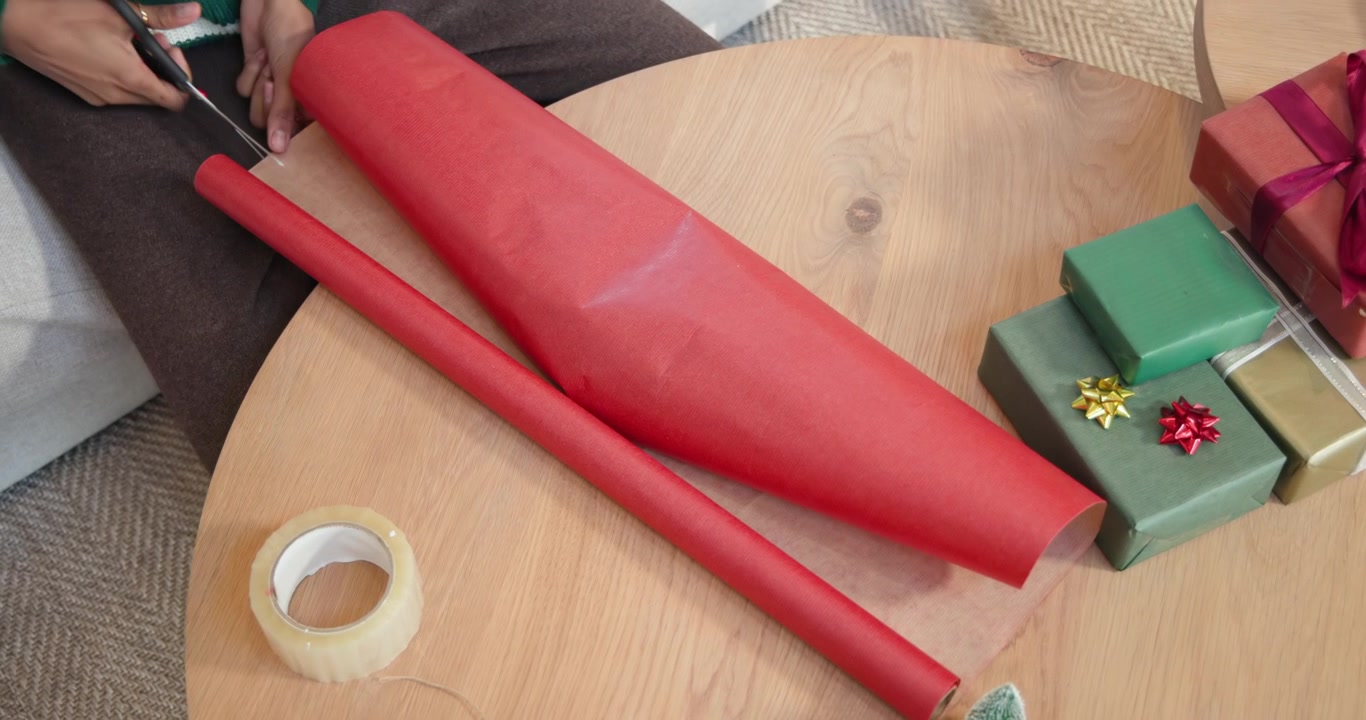 Hands are placing box on kraft paper on table, unrolling red paper, trimming excess for gifts