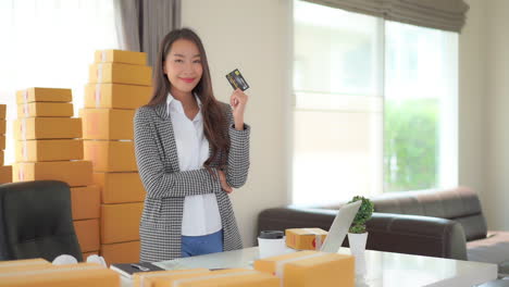 static shot of a cute asian lady standing in an office surrounded by boxes while holding a credit card in one hand and doing a thumbs up in the other hand