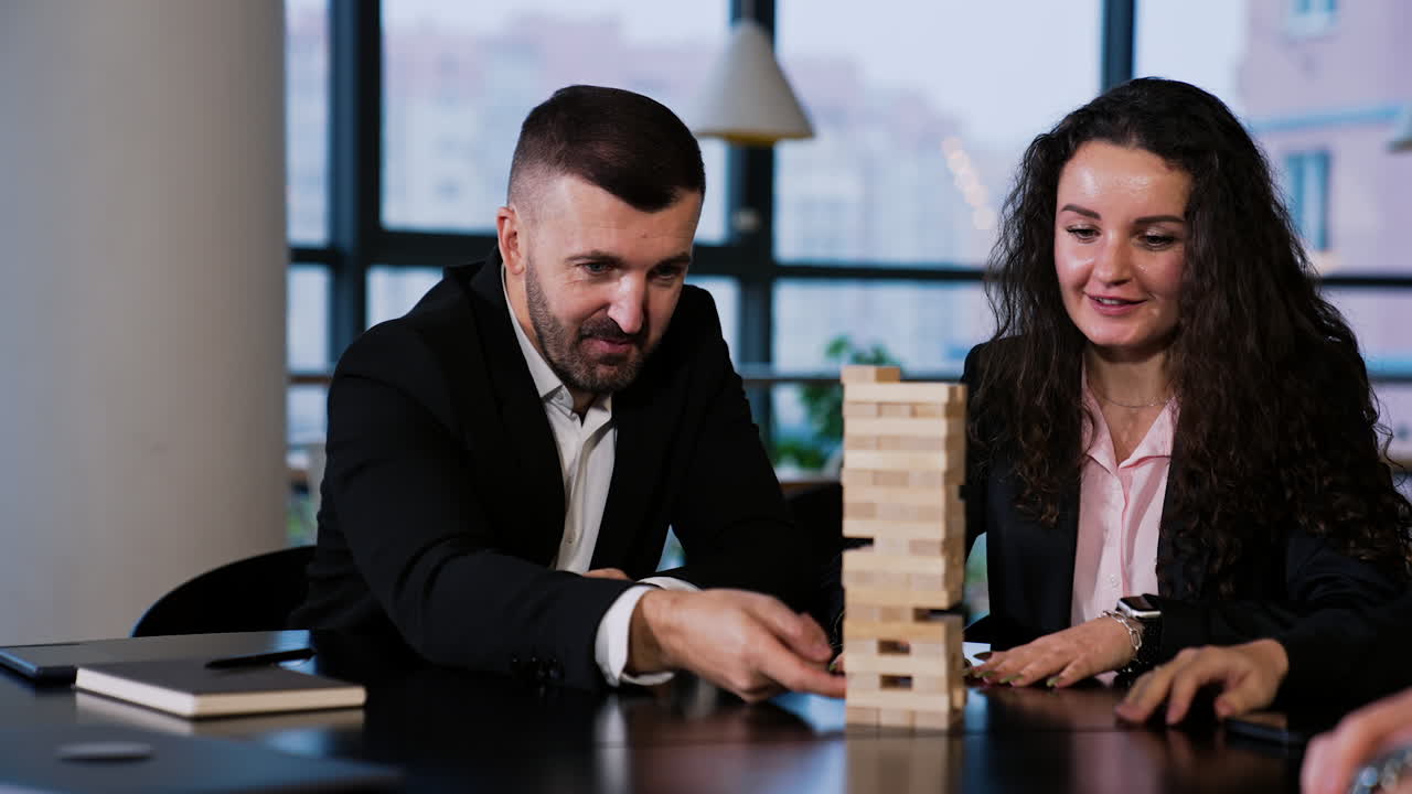 Adult people sitting at table play table games. Bearded smiling man tries to move the wooden brick in little tower of jenga.