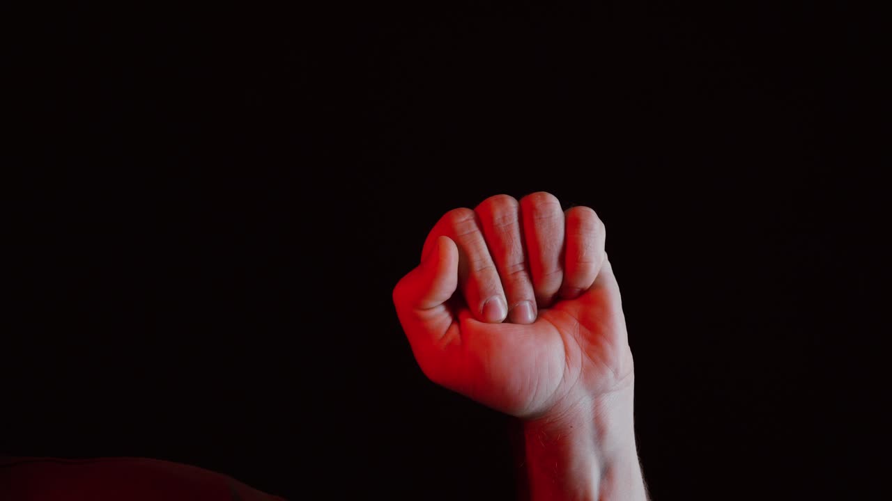 A close-up of a man’s hand counting to three with his fingers under dramatic red lighting against a dark background, symbolizing tension, countdown, or suspense