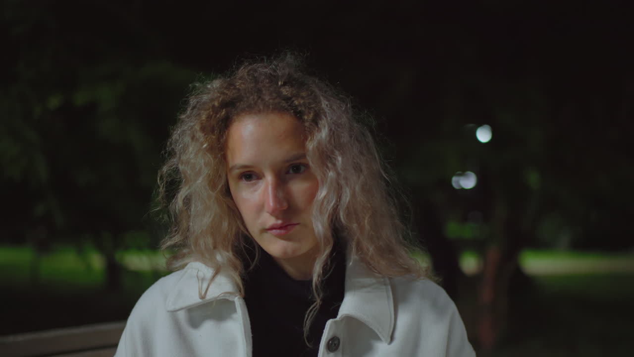 Young woman with curly hair in white coat lifts head thoughtfully in dim park light at night, seated in peaceful outdoor space with blurred greenery in background and gentle expression on her face