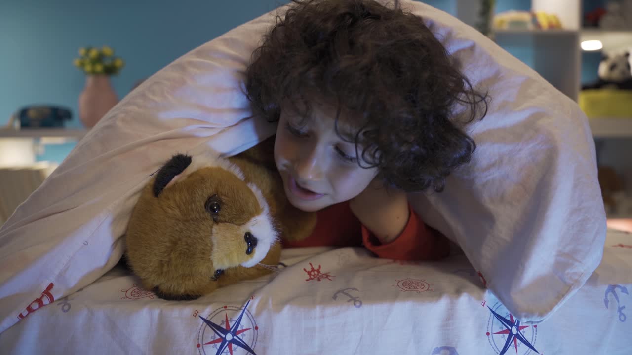 귀여운 소년이 장난감 개와 함께 침대  ⁇ 에 숨어 놀고 있습니다.