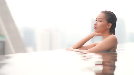 young asian wooman standing sumberged in the swimming pool, looking happy and relaxing admiring the view of the city