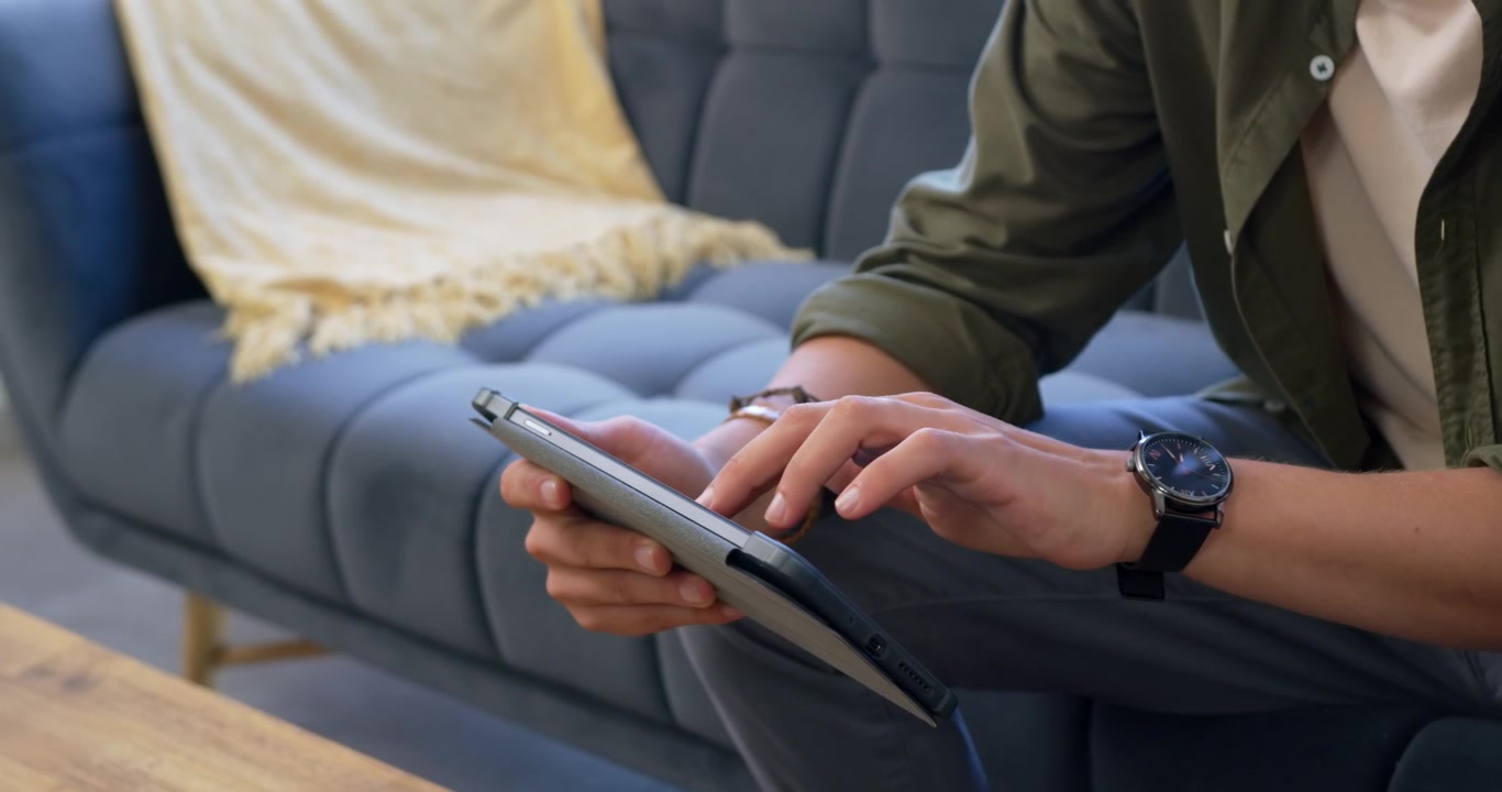 Uomo adulto seduto su un divano capitonné, con la mano sinistra che sorregge un tablet collegato via cavo, che tocca per leggere.