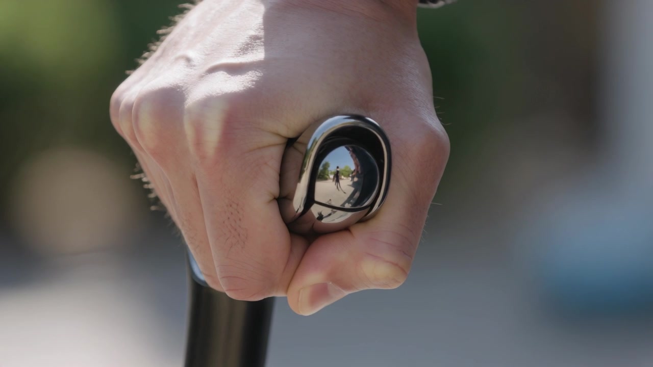 Mano adulta sulla barra che regola il cambio anteriore, con il riflesso del tappo terminale che cambia a causa di una persona sul marciapiede.