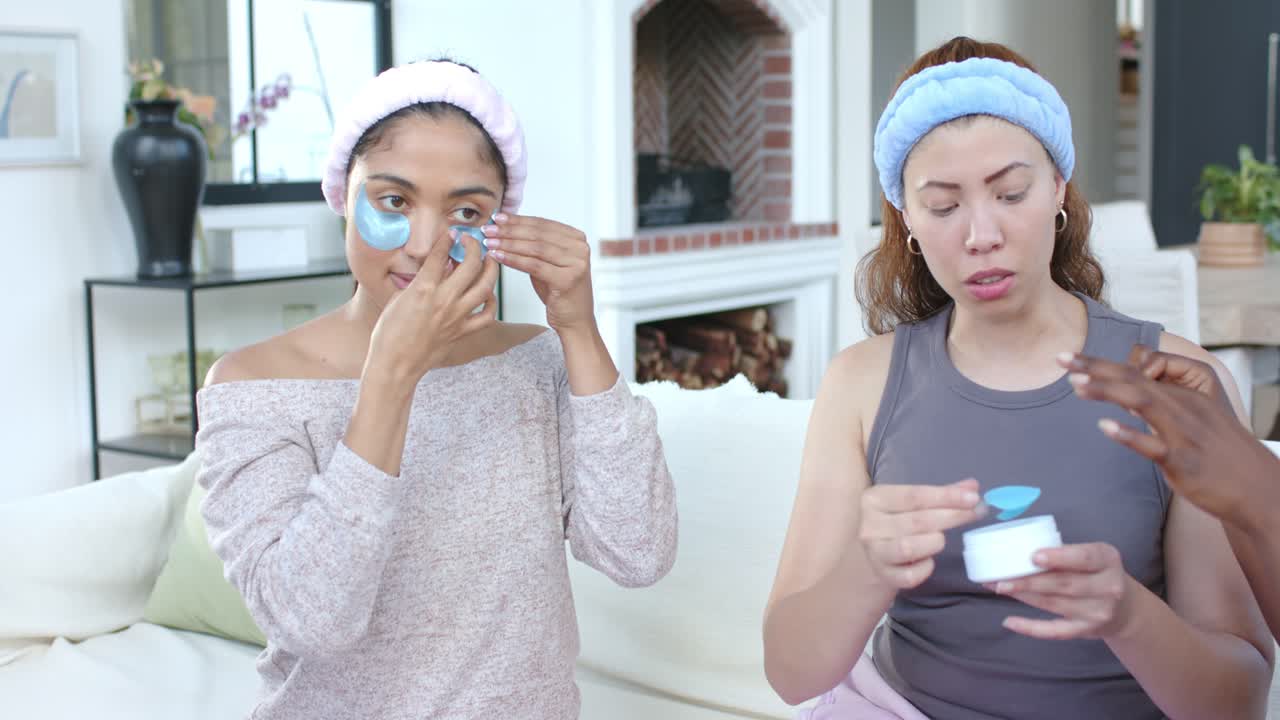 Female friends on sofa applying blue under-eye patches peeling backing, sharing cream for skincare