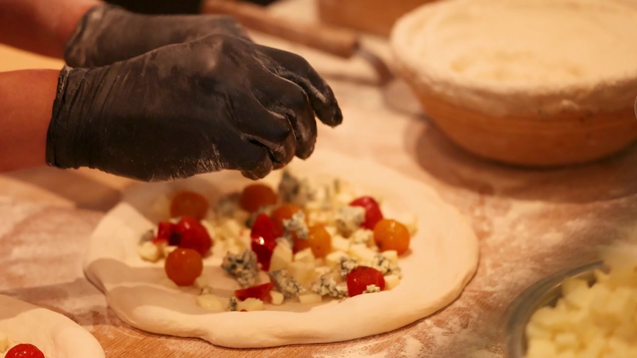 Primo piano di uno chef che posiziona pomodorini e formaggio erborinato su impasto crudo per pizza.