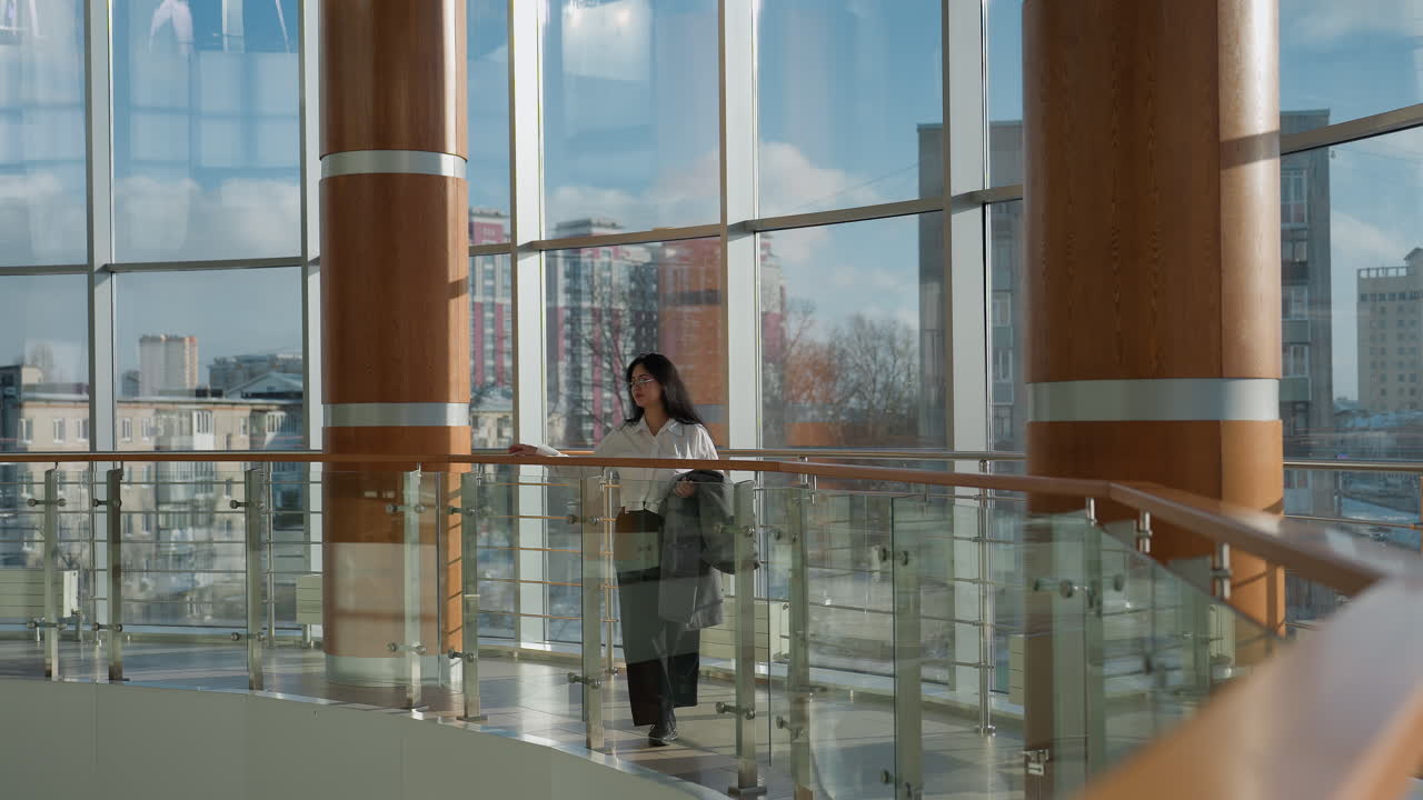 Stylish professional woman in white shirt and glasses walking along mall gallery lined with glass and wooden railings, city skyline visible through tall windows on bright sunny day