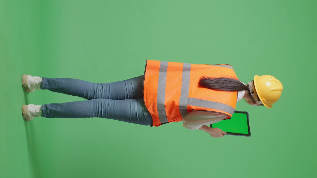 Full Body Back View Of Asian Female Engineer With Safety Helmet Working On A Green Screen Tablet And Looking Around While Standing In The Green Screen Background Studio
