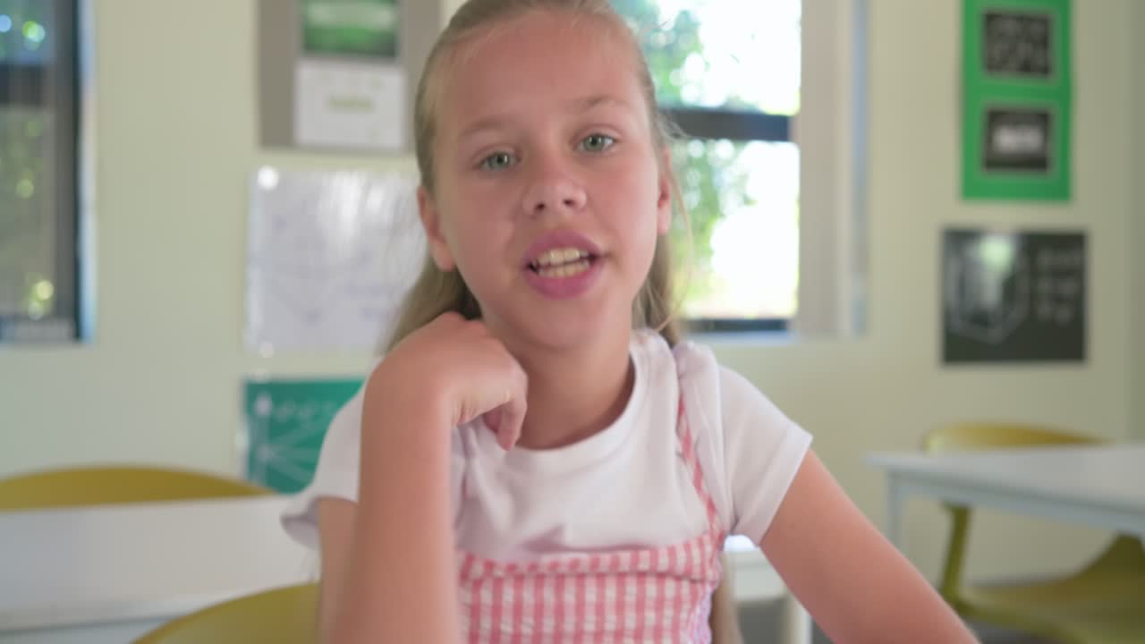 Teenager-Mädchen lächelt und spricht dann in der Klasse am Schreibtisch in die Kamera und hält eine Präsentation in einem rosa Schürzenkleid