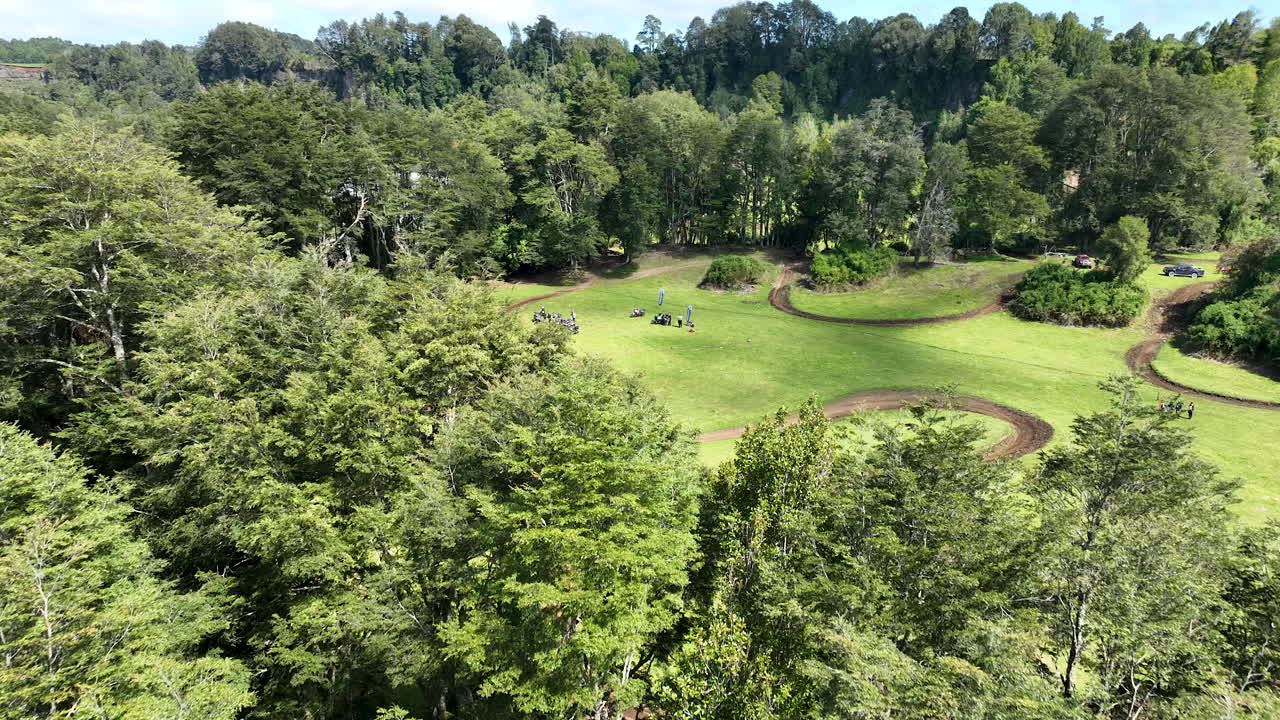 Vuelo a baja altitud pasando cerca de las copas de los árboles para revelar una zona de organización de un evento de motocicletas con personas y vehículos en un campo verde en Chile