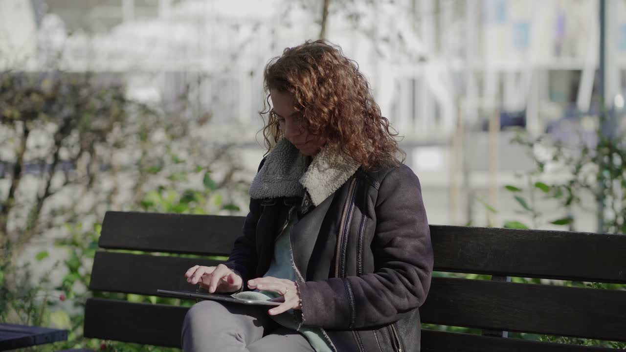 Concentrated curly woman typing on tablet while sitting on wooden bench