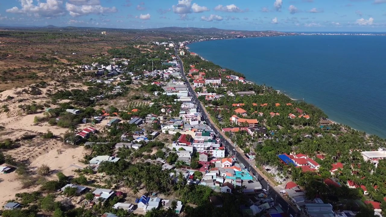 Riprese aeree con drone di Mui Ne, Vietnam, che mostrano una strada costiera circondata da hotel, resort, edifici residenziali e vegetazione tropicale, con l'oceano blu e la spiaggia visibili nelle vicinanze