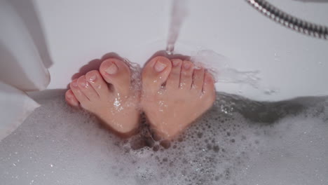 woman holds feet under running water taking bath at home. lady massages tired legs with liquid flow in bathroom. hygiene and relaxation in bath