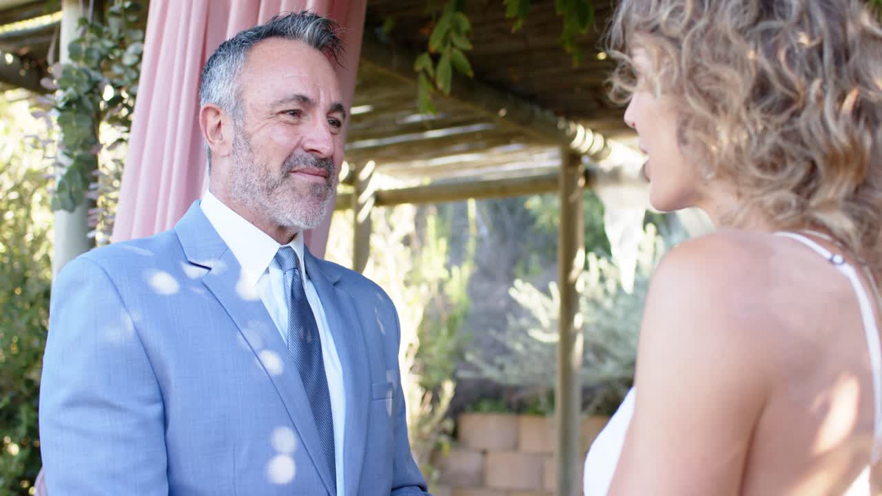 Pareja intercambiando anillos después de los votos bajo un arco, hombre de traje azul colocando el anillo en la mujer de vestido blanco.