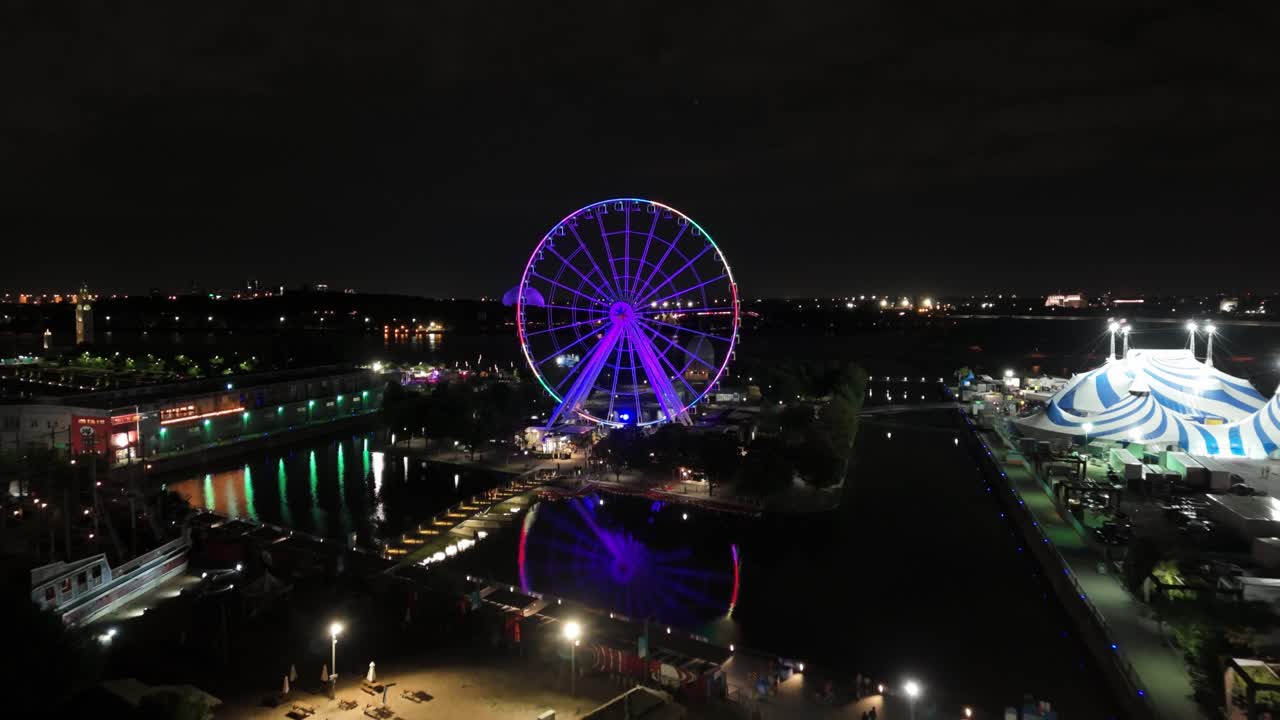 Drone footage of a Ferris wheel glowing with blue and white lights at night, located by the waterfront with city lights in the background and a striped circus tent nearby