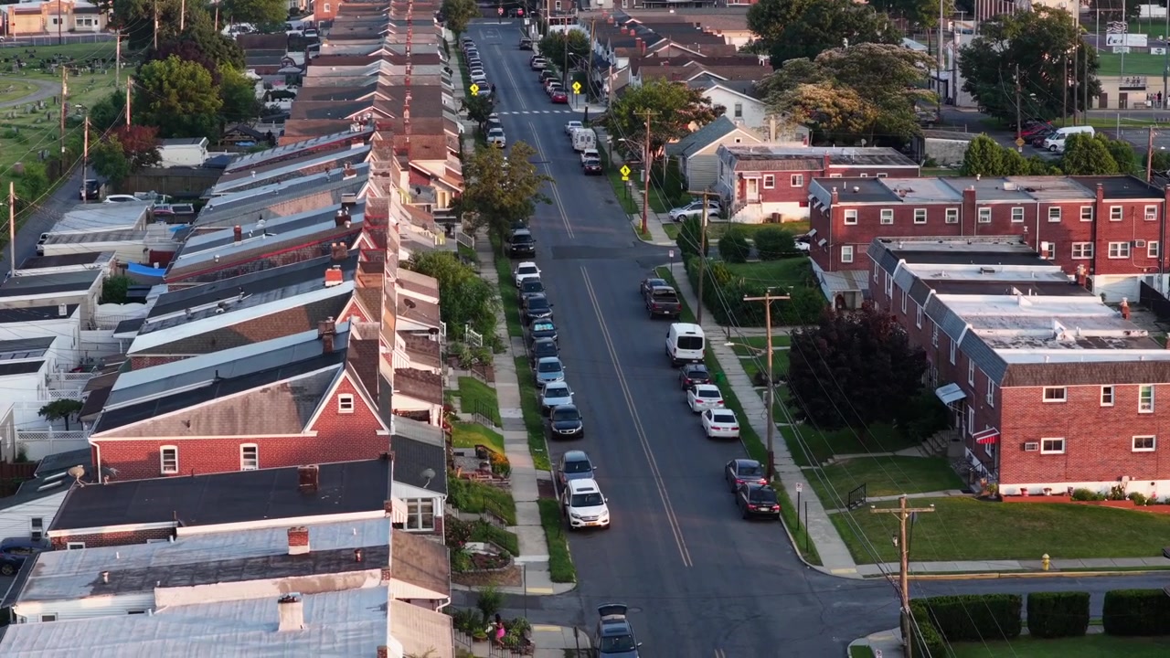 Case a schiera con tetti in metallo fiancheggiano una strada diagonale ad Allentown, Pennsylvania, con case a schiera in mattoni e prati verdi che si estendono nel quartiere mentre la luce del tramonto riscalda i tetti. Veduta aerea con drone