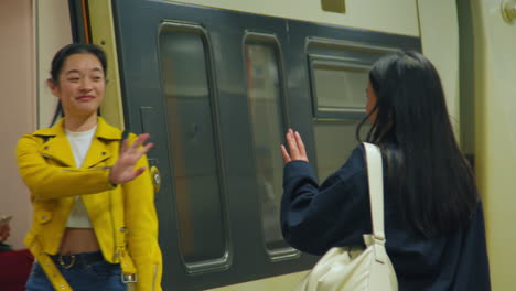 Two-Young-Female-Friends-Waving-As-They-Say-Goodbye-On-Underground-Train-Station-Platform