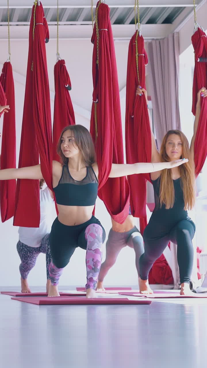 concentrated slim ladies practice warrior asana holding red fly yoga hammocks above heads in modern gym hall