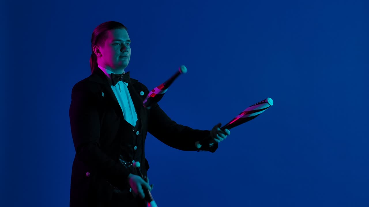 стильный шоумен жонглирует булавками в темной студии. человек мастерски бросает и ловит булавки, концентрируясь на них. голубой экран, хроматическая клавиатура.