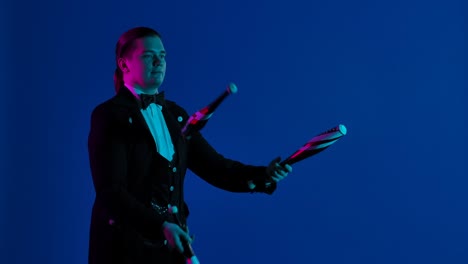 un elegante showman hace malabarismos con alfileres en un estudio oscuro. el hombre lanza y atrapa los alfileres con maestría concentrándose en ellos. pantalla azul, tecla de croma. primer plano. cámara lenta