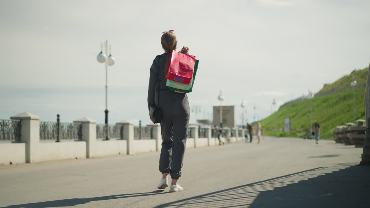 la señora disminuye la velocidad, ajustando las bolsas de compras en su hombro mientras camina cerca de las escaleras del puente, el fondo presenta una vista borrosa de la gente en la distancia, una colina y un paisaje pintoresco