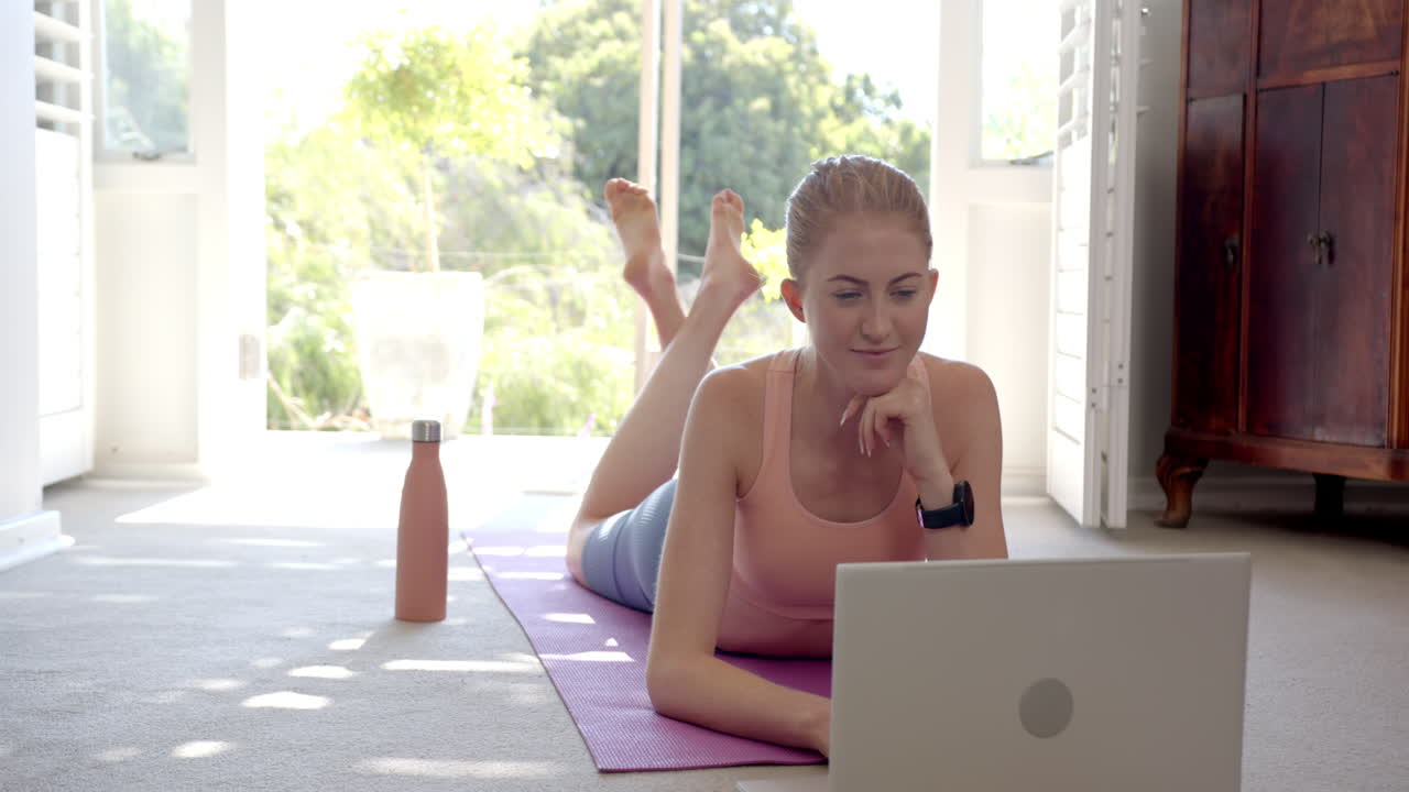 Lying on yoga mat, woman using laptop and water bottle at home