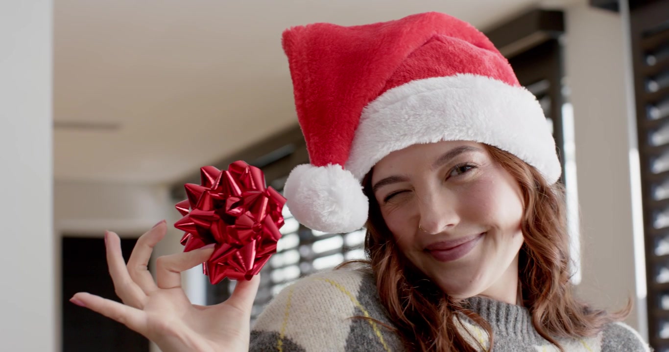 Adult woman in Santa hat holding red bow over eye, peeking, presenting for holiday at window