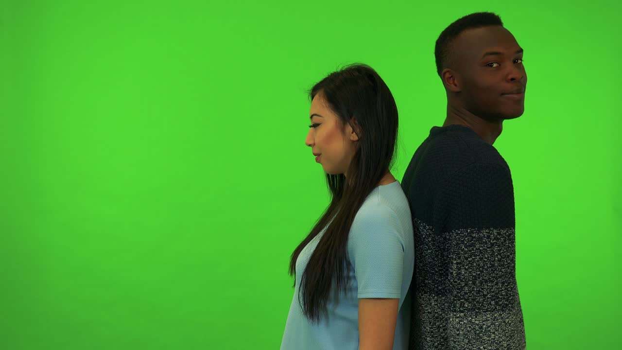 Uma jovem mulher asiática e um jovem homem negro ficam de costas um para o outro e se viram para a câmera com um sorriso - estúdio de tela verde.