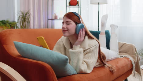 Mujer-Joven-Feliz-Con-Auriculares-Inalámbricos-Relajándose-Tumbada-En-El-Sofá-De-Casa-Escuchando-Su-Música-Favorita