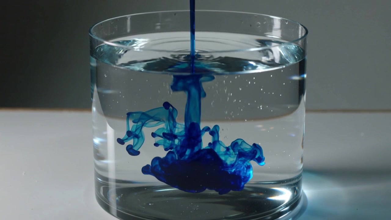 Un dispenser rilascia sopra un bicchiere su un tavolo bianco, versando colorante blu nell'acqua, formando pennacchi che affondano.