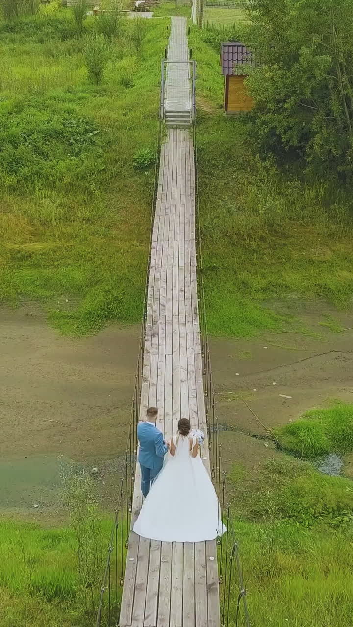 young just married couple walks along rustic wooden bridge over stream with green banks upper backside view