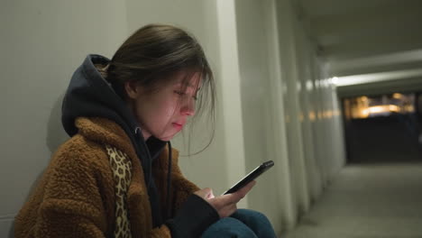 una toma de primer plano de una chica con un abrigo marrón, sentada inclinada en un túnel subterráneo, revisando su teléfono con una expresión llorosa. la escena evoca una profunda sensación de tristeza, soledad