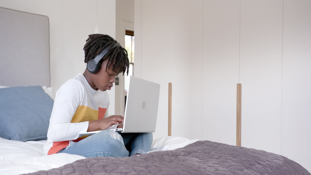 African american boy wearing headphones sitting on bed using laptop at home, slow motion, copy space