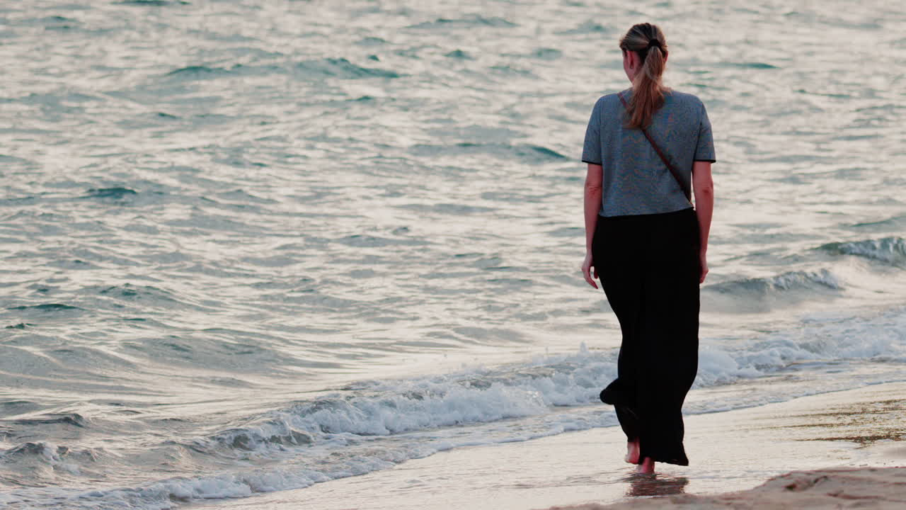 A woman walks barefoot along the beach, waves gently touching her feet