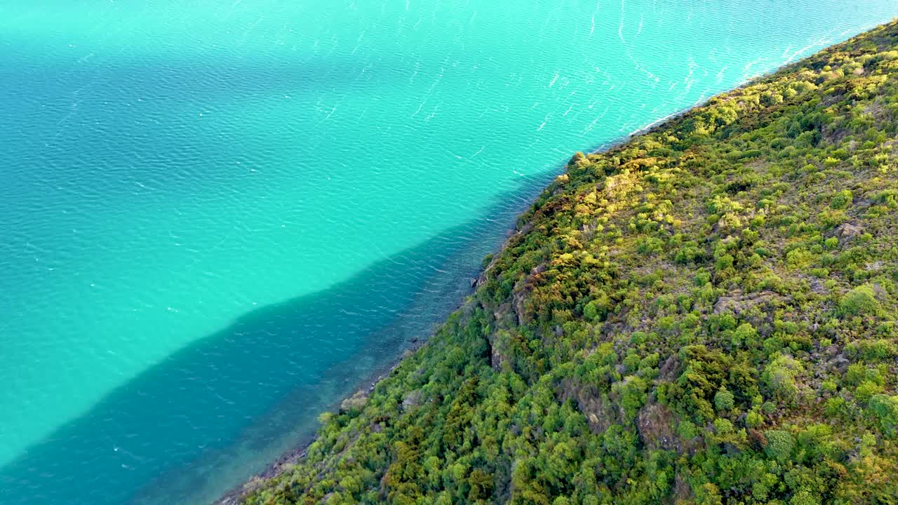 Drone glides above lush hillside and vibrant blue lake, bright daylight, smooth lateral movement