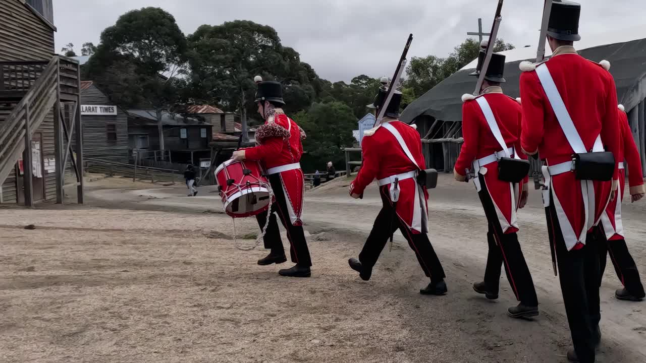 A group of soldiers in red uniforms march past wooden structures on a gravel path.
