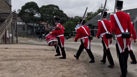 a group of soldiers in red uniforms march past wooden structures on a gravel path.