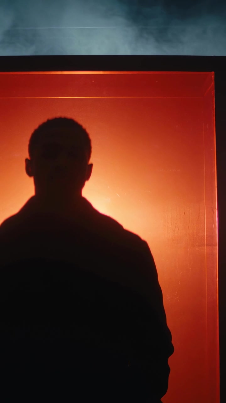 Vertical video: Tilting man silhouette moving head in studio with framed orange panel, haze, jacket