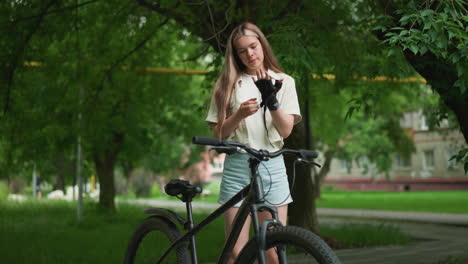 young adult adjusts black glove while standing beside her bicycle on a paved path, background showcases lush greenery with a blurred view of building