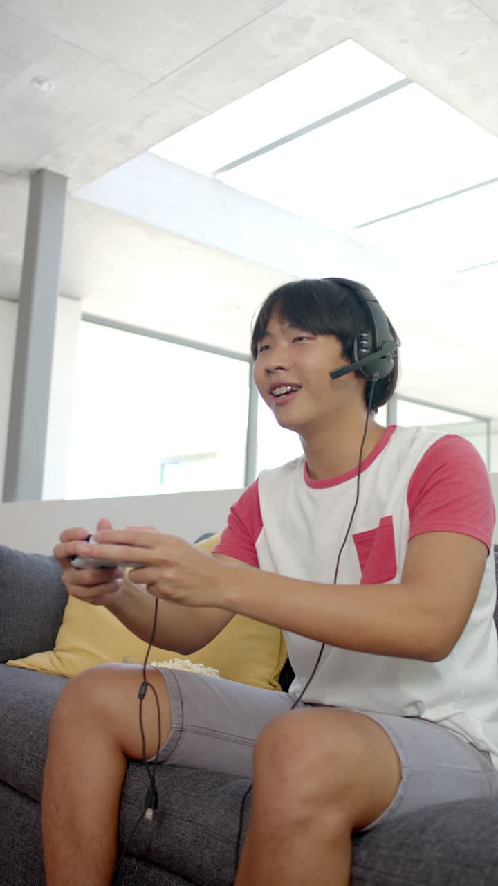 Vertical video: Playing video game, Asian teenage boy wearing headset holding controller, sitting on