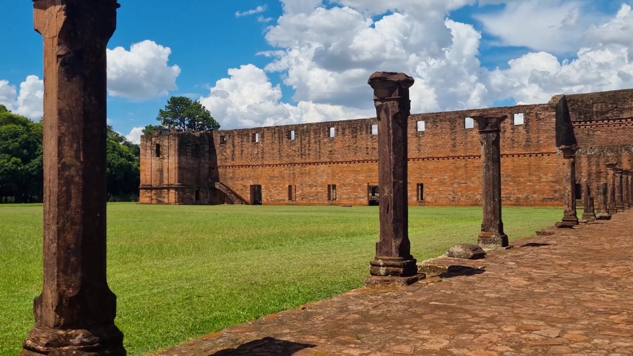 Rovine illuminate dal sole della missione gesuita Jesús de Tavarangüé in Paraguay si ergono sotto un cielo azzurro limpido, con erba verde che si estende di fronte alla storica struttura in mattoni, a testimonianza della storia missionaria