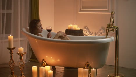 woman lying in bubble bath with the light from the candles to relax and drink wine. romantic relaxing aromatherapy atmosphere. bathroom with candles.