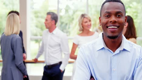 Male-business-executive-smiling-at-camera-while-colleagues-interacting-in-background